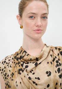 Blusa drapeada beige con un patrón de leopardo, que presenta una textura suave y un escote enrollado. Pendientes de gota en tono dorado complementan el diseño.