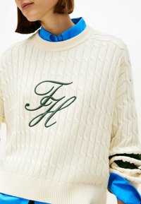 Pulover tricotat de culoare crem, cu un model texturat, având un logo brodat verde pe partea stângă a pieptului și un tiv elastic.