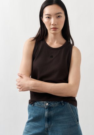 Joven mujer con cabello largo y oscuro lleva una camiseta negra sin mangas y vaqueros azules, de pie con los brazos cruzados frente a un fondo blanco liso.