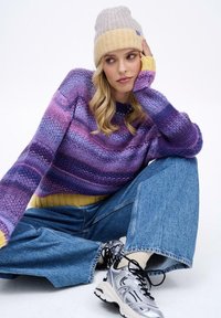 Fialovo-ružový pruhovaný pletený sveter so žltým lemom, v kombinácii s svetlomodrými širokými džínsami a striebornými teniskami, doplnený svetlou čiapkou.