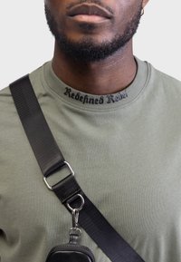 Olivinės spalvos medvilninis marškinėliai su juodu užrašu "Redefined Rebel" ant kaklo, derinami su juodu reguliuojamu dirželiu ir kišenėle.