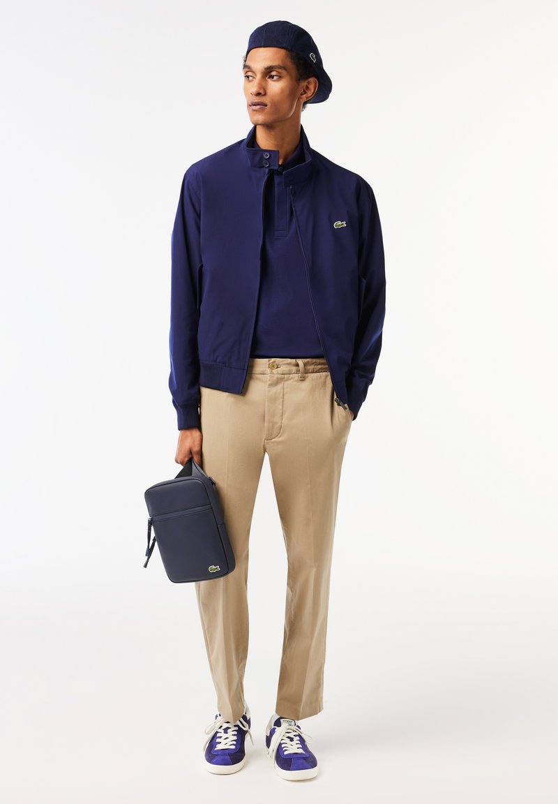 Veste zippée navy sur un polo navy, pantalon beige et baskets bleues. Un sac navy avec un logo vert est tenu dans une main.