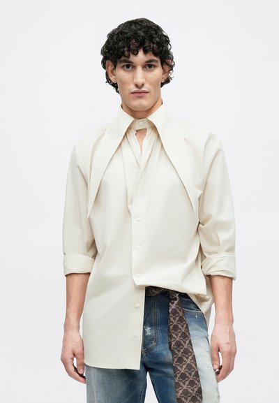 Beige long-sleeve shirt with unique collar design, featuring a button front and relaxed sleeves, paired with distressed denim and patterned accents.