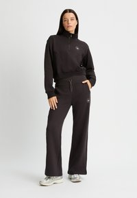 Sudadera negra con cremallera y cuello alto, con detalles de costura y un pequeño logo. Combinada con pantalones de chándal negros de corte holgado y con bolsillos.