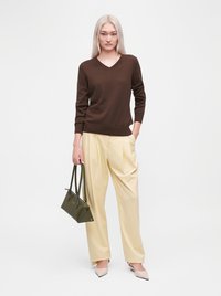 Femme aux longs cheveux blonds porte un pull marron col en V, un pantalon large crème, des chaussures beiges et tient un sac à main vert foncé sur un fond uni.