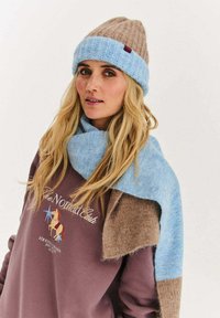 Pletená čepice v hnědé a světle modré barvě s ohrnutým lemem; odpovídající modrá šála přehozená přes hnědou a fialovou mikinu s logem.