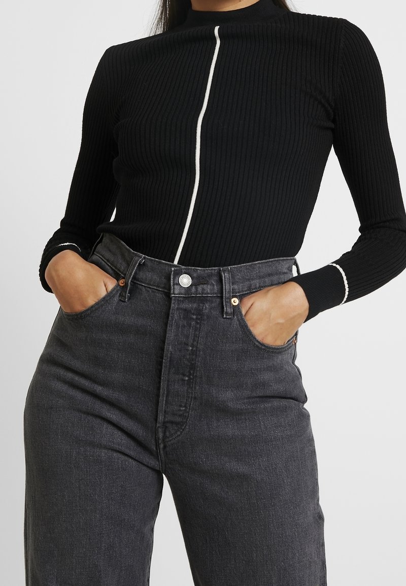 Haut noir à manches longues côtelé avec un col montant et un détail de fermeture éclair blanche contrastante. Associé à un jean en denim gris foncé taille haute.