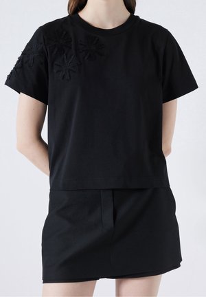 T-shirt in cotone nero con maniche corte, caratterizzato da fiori in tessuto nero appuntati su una spalla. Taglio semplice e dritto con scollatura rotonda.