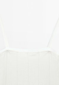 Top en lin blanc à fines bretelles, présentant une surface texturée et des coutures verticales. L'encolure est bordée d'une bordure subtile.