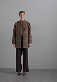 Bruine en beige geblokte jas met twee borstzakken, gedragen over een beige top en gecombineerd met een donkere, wijde broek en zwarte schoenen.