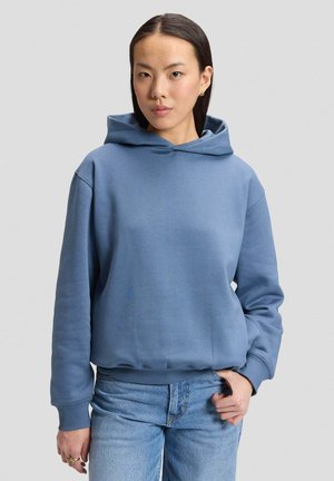 Junge Frau mit glatten dunklen Haaren trägt einen blauen Hoodie und hellblaue Jeans, steht mit einer Hand in der Tasche vor einem schlichten Hintergrund.