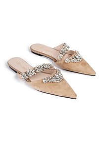 Beige mocka spetsskor med öppen tå, lätt att ta på, med kristalldekorationer på remmarna och foten, uppställda på vit bakgrund.