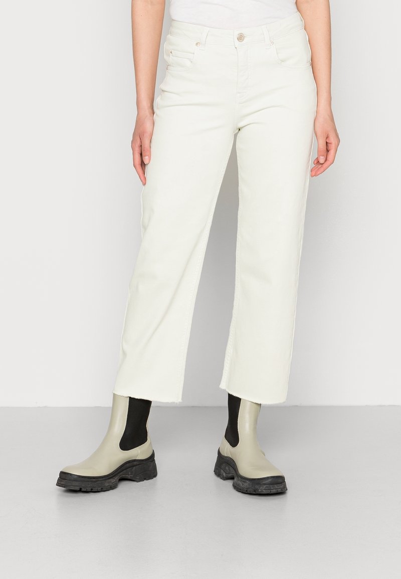 Jean blanc taille haute, court, avec une coupe droite, des ourlets légèrement effilochés, et des poches devant et derrière. Associé à des bottines grises.