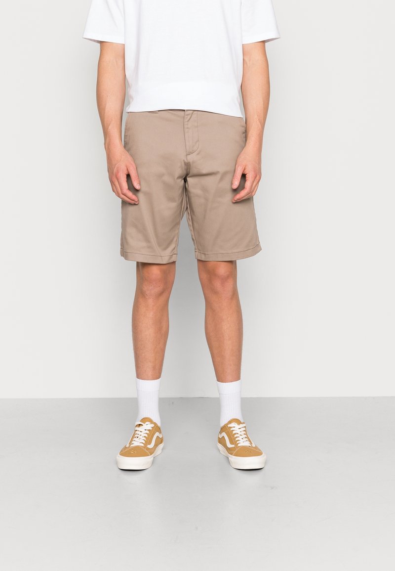 Shorts beiges avec une coupe droite, fabriqués en tissu léger. Associés à des chaussettes blanches mi-mollet et des baskets beiges avec une rayure blanche.