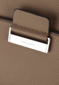 Brązowa skórzana tekstura z srebrnym akcentem z napisem "GERRY WEBER". Materiał ma lekko ziarnistą fakturę.