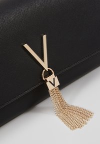 Musta kädessä pidettävä laukku teksturoidulla pinnalla, kultainen "V"-logo ja kiinnitetty kultainen ketjutupsu. Minimalistinen muotoilu ja tyylikäs muoto.