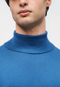 Jeune homme portant un pull à col roulé bleu ajusté, montrant le cou et le bas du visage sur un fond clair uni.