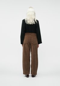 Pikend blondide juustega inimene kannab musta pika varrukaga särki ja kõrge vöökohaga pruune puuvillaseid laiad püksid, seistes seljaga poole.