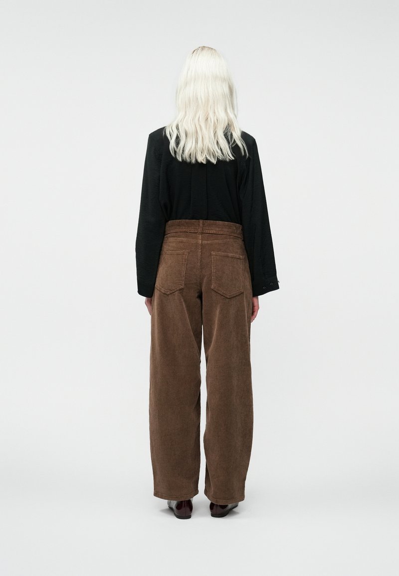 Une personne avec de longs cheveux blonds porte une chemise noire à manches longues et un pantalon large en velours côtelé marron taille haute, se tenant de dos.