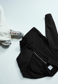 Crna jakna sa patentnim zatvaračem od teksturirane tkanine, savijena na svijetloj podlozi uz sivi i bijeli sportski tenis.
