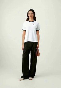 Camiseta blanca de manga corta, con cuello negro acanalado y logo de "Coach", combinada con pantalones negros sueltos y zapatos de cama beige.