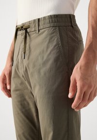 Man in olijfgroene trekkoordbroek en een witte geribde shirt, met zichtbare voor- en zijzakken en een relaxte pasvorm.