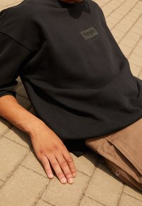 Felpa nera con logo rettangolare, materiale testurizzato, vestibilità comoda. Mani appoggiate su pantaloni in tessuto marrone chiaro, superficie piatta sottostante.