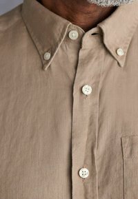 Beige overhemddie gemaakt is van textiel met textuur, voorzien van een puntige kraag, witte knopen en een enkele borstzak.