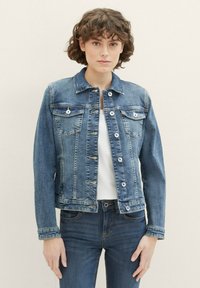 Giacca di jeans blu medio con colletto, bottoni frontali, due tasche sul petto e maniche lunghe. Presenta un design aderente e linee pulite.
