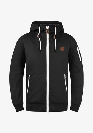 Svart lett jakke med glidelås foran, justerbar hette, hvite detaljer, sidelommer og et brunt skinnlogo merke.