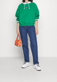 Sudadera verde con cordones blancos y puños a rayas, jeans azules, zapatillas blancas y un bolso a cuadros naranja.