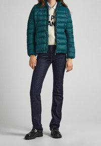 Teal puffermantel med horisontell quiltning, dragkedjestängning och ribbade ärmmuddar över en krämfärgad tröja med svart text, parat med mörk jeans och svarta skor.