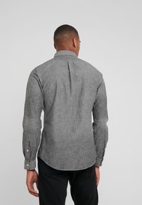 Camicia in chambray grigio a maniche lunghe con colletto classico e polsini abbottonati, caratterizzata da una piega sul retro e orlo curvo.