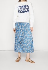 Nainen, joka pukeutuu valkoiseen huppariin, jossa on teksti "Sandro 1984 Paris", siniseen kirjailtuun laskostettuun midi-hameseen ja kultaisiin litteisiin sandaaleihin, seisoo.