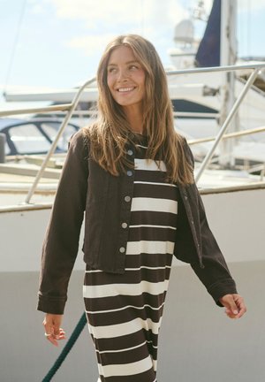 Ung kvinde med langt hår iført en sort og hvid striped kjole og en sort jakke, smilende udendørs nær både og gelændere.