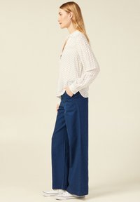 Blusa bianca a maniche lunghe con un piccolo motivo geometrico, abbinata a pantaloni blu a gamba larga. Il modello è in posa di profilo con le mani nelle tasche.