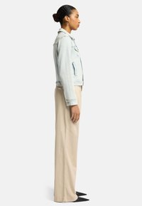 Lysblå denimjakke med et klassisk design, der har krave og sidelommer, båret med beige bukser og sorte spidse sko.