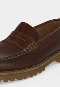 Mocassin marron en cuir avec détails cousus, bride penny et semelle épaisse à crampons pour une meilleure adhérence.