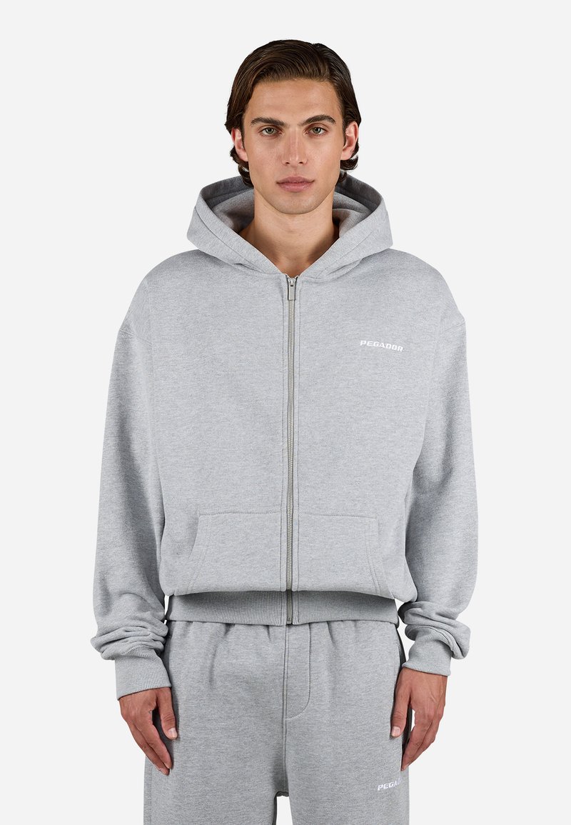 Gray zip-up hoodie with a front pocket, cotton blend fabric, ribbed cuffs, and a small white logo on the chest. Styled with matching sweatpants.
