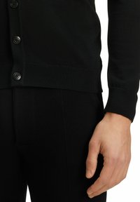 Cardigan nero con una fila singola di bottoni neri, caratterizzato da una texture a coste all'orlo e ai polsini. Primo piano di una mano appoggiata su una gamba dei pantaloni.