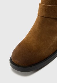 Pruun nubukist ankle'i saabas ümarate varvaste ja musta kummitaldaga. Omab riba ümber pahkluu kindla istuvuse tagamiseks. Sile tekstuur.