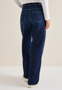 Vaqueros de mezclilla azul oscuro con un corte recto. Cuenta con dos bolsillos traseros y costuras blancas en contraste. Llevado con una blusa de manga larga blanca.