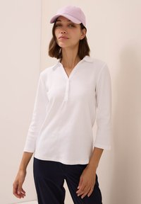 Polo shirt bianco a maniche lunghe con colletto a alette, dotato di una chiusura a bottoni e realizzato in morbido cotone, abbinato a un cappellino di un leggero colore viola.