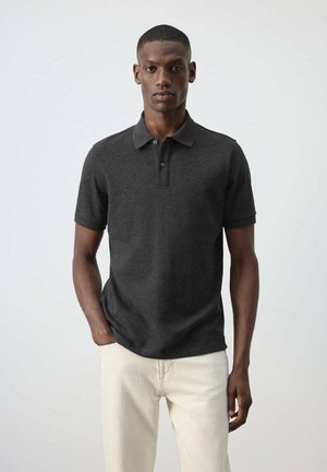 Jeune homme debout face à l'avant, portant un polo à manches courtes gris foncé et un pantalon beige clair avec une main dans la poche.