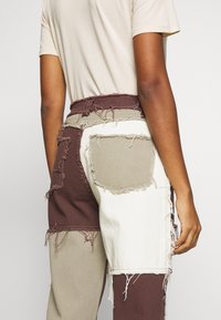 Person wearing color-blocked patchwork pants in brown, beige, and white with frayed edges, paired with a beige short-sleeve top.