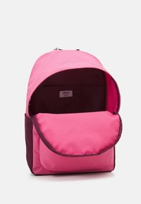 Rosa tygryggsäck med klaffstängning, mörklila botten och stort öppningsfack. Insidan har en etikett och en slät textur.