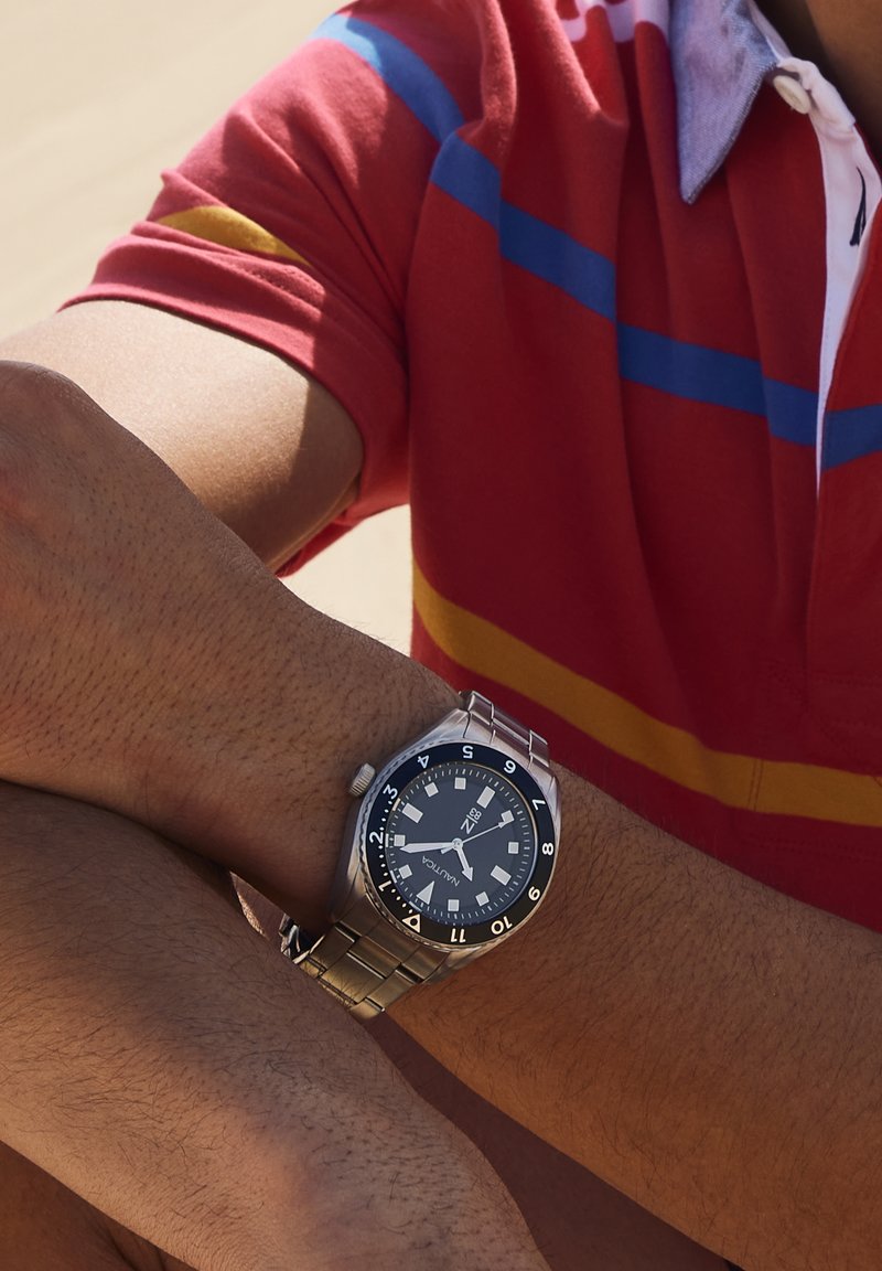 Hombre con un reloj de pulsera de metal plateado con esfera negra en su muñeca izquierda, vestido con una camiseta tipo polo roja con rayas azules y amarillas.