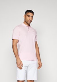 Ljusrosa polo-tröja med krage, korta ärmar och tre knappar. Har en liten grå logotyp på vänster bröst. Bärs med vita shorts.