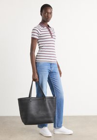 Svart lær tote bag med en strukturert form og lange håndtak, kombinert med en stripete polo-skjorte og blå jeans. Modellen står mot en nøytral bakgrunn.