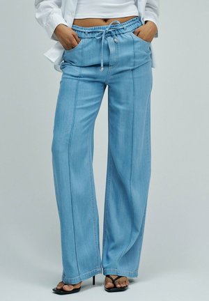 Femme portant un jean large bleu clair avec poches avant et taille à cordon, associé à des sandales noires à talons, un top blanc court et une chemise ouverte.